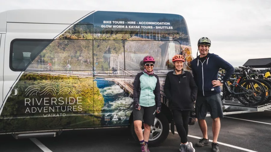 Group of cyclists in helmets posing beside a Riverside Adventures Waikato shuttle van and bike trailer