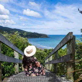 Woman in a floral dress and sunhat sitting on a wooden stairway overlooking Raglan Beach, Waikato, New Zealand.