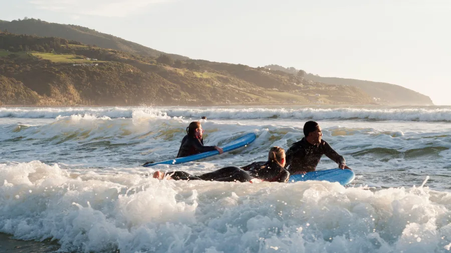 Surfers paddling through waves at Raglan Beach in Waikato, New Zealand, with green hills in the background.