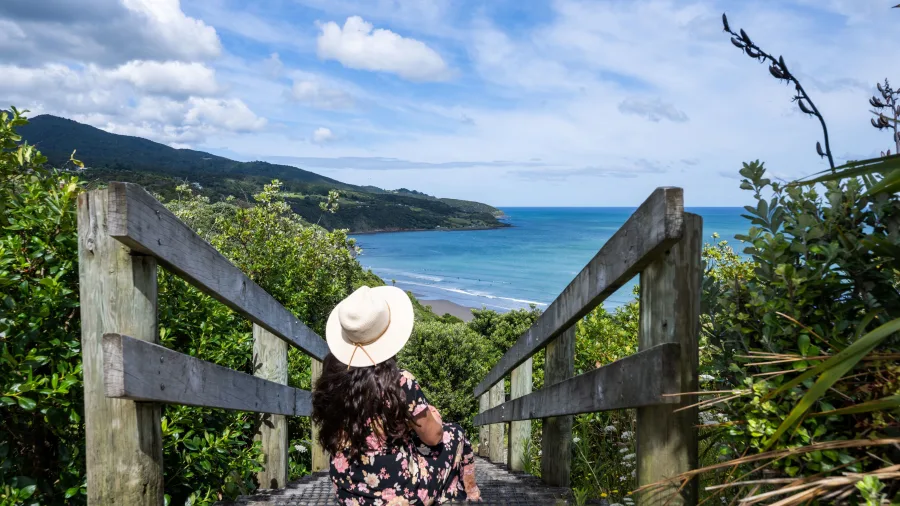 Woman in a floral dress and sunhat sitting on a wooden stairway overlooking Raglan Beach, Waikato, New Zealand.