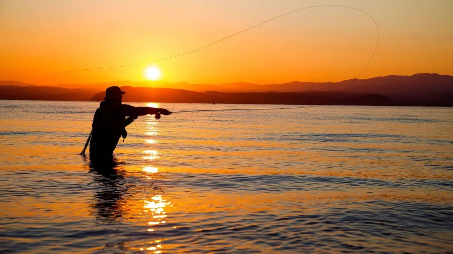 Silhouette of an angler fly fishing at Waitahanui on Great Lake Taupo during a vivid orange sunrise in New Zealand.