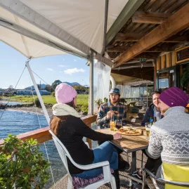Friends relaxing with drinks and freshly made wood-fired pizza at 2 Mile Bay Sailing Centre on the shores of Lake Taupō, New Zealand.