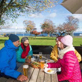 Friends enjoying drinks and freshly made pizza at Dixie Browns restaurant on the lakefront in Taupō, New Zealand, with views of Lake Taupō.