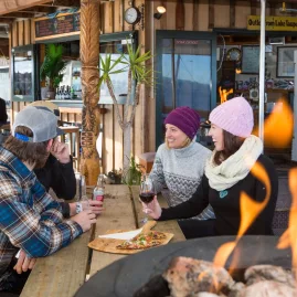 Friends enjoying drinks and pizza by the fire at Mile Bay Watersports Centre Bar and Café in Taupō, New Zealand.