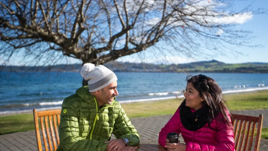 Couple enjoying takeaway coffee at The Steaming Bean on Lake Terrace, Taupō, with lake views and a backdrop of rolling hills in New Zealand.