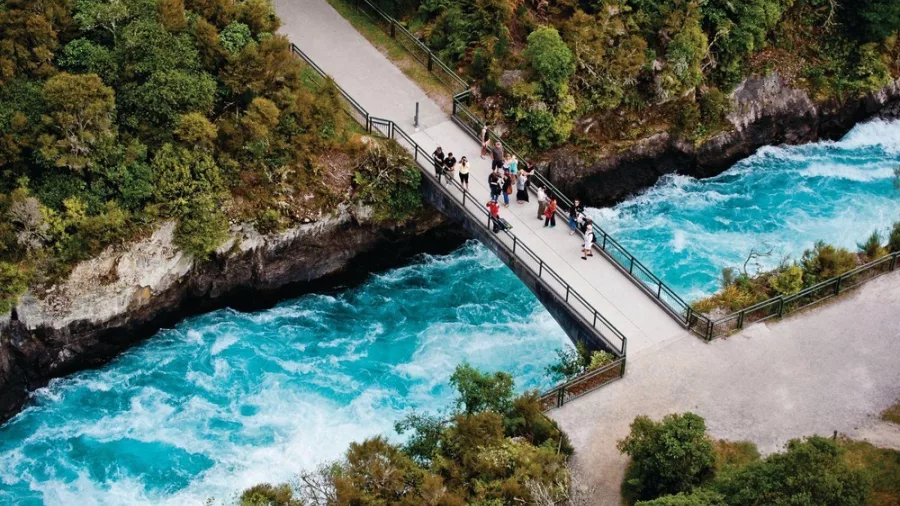 Visitors standing on the pedestrian bridge over the vibrant blue waters of Huka Falls, surrounded by lush greenery near Lake Taupō, New Zealand