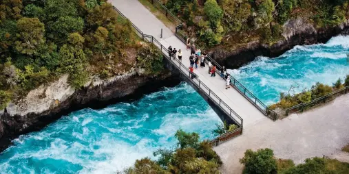 Visitors standing on the pedestrian bridge over the vibrant blue waters of Huka Falls, surrounded by lush greenery near Lake Taupō, New Zealand