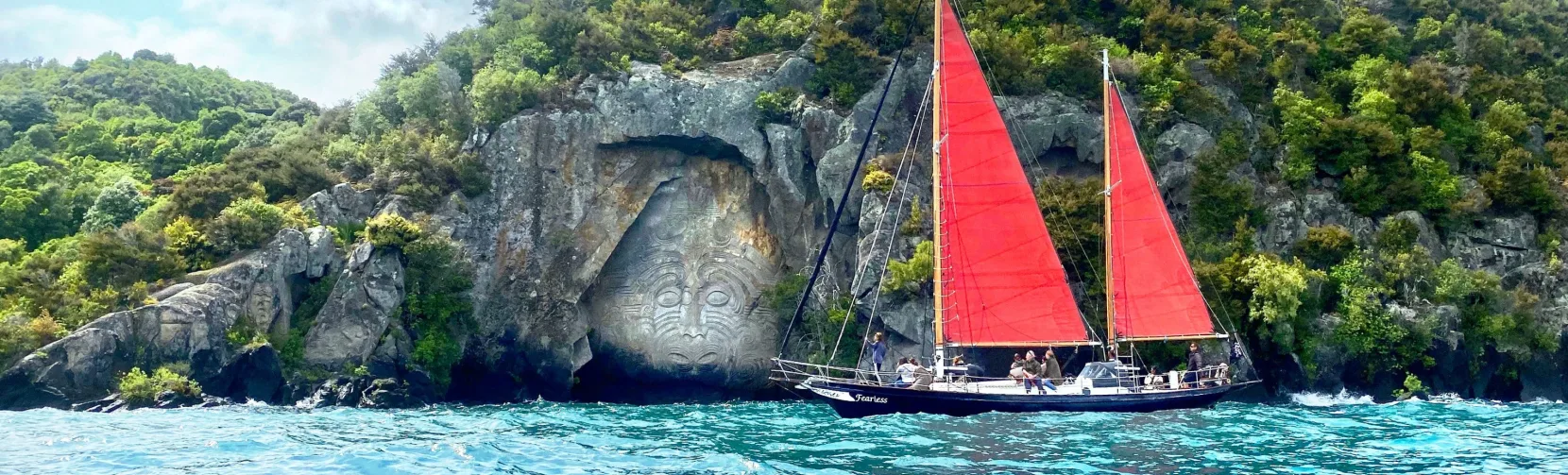 Red-sailed yacht cruising past the Māori rock carvings at Mine Bay on Lake Taupō