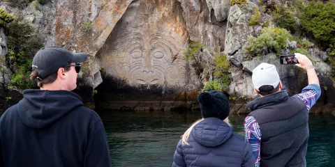Tourists taking photos of the Ngatoroirangi Mine Bay Maori Rock Carvings from a boat on Lake Taupō