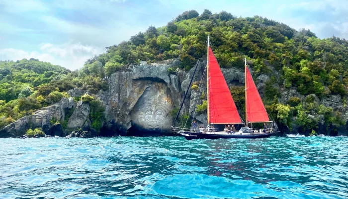 Red-sailed yacht cruising past the Māori rock carvings at Mine Bay on Lake Taupō