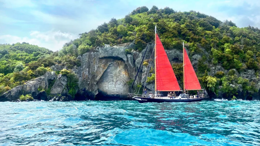 Red-sailed yacht cruising past the Māori rock carvings at Mine Bay on Lake Taupō