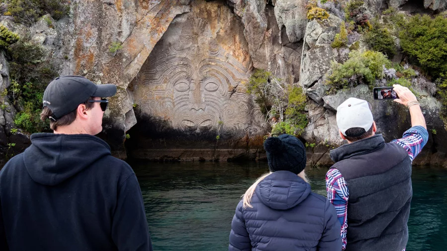 Tourists taking photos of the Ngatoroirangi Mine Bay Maori Rock Carvings from a boat on Lake Taupō
