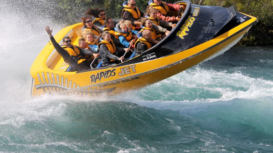 Passengers on a yellow Rapids Jet boat riding through white-water rapids near Taupō, New Zealand, with water splashing high around them.