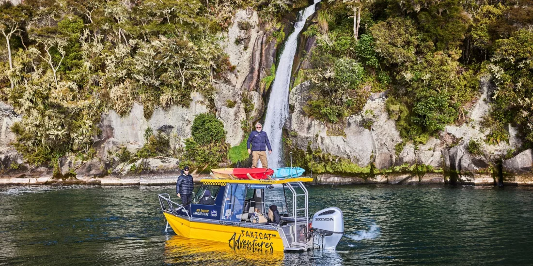 Yellow Taxicat boat floating near a lakeside waterfall with people standing on board in Taupō