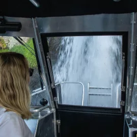 Woman inside a boat looking through the rear door at a waterfall pouring into Lake Taupō
