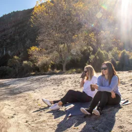 Two women sitting on Waihora Beach enjoying drinks with sun rays and native bush behind them in Taupō