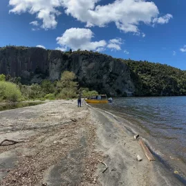 Person walking from a yellow boat on Waihora Beach with cliffs and native bush in Taupō