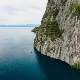Aerial view of steep cliff face meeting the deep blue waters of Lake Taupō with a small yellow boat below