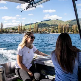 Two women enjoying a lake boat ride with scenic hills and trees in the background near Waitomo