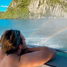Man on a boat admiring a rainbow formed by water spray near cliffside on a lake tour in Waitomo