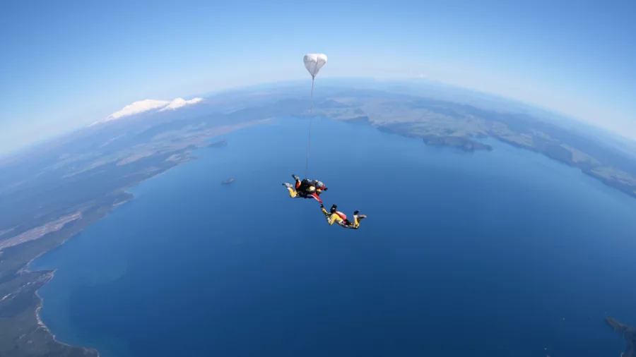 Tandem skydivers in freefall above the deep blue waters of Lake Taupō, New Zealand, with a parachute deploying.