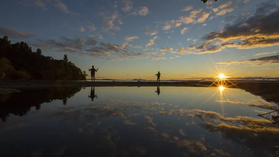 Silhouettes of two anglers trout fishing at the Kuratau River mouth in Turangi during sunset, with reflections on the water at Lake Taupō, New Zealand.