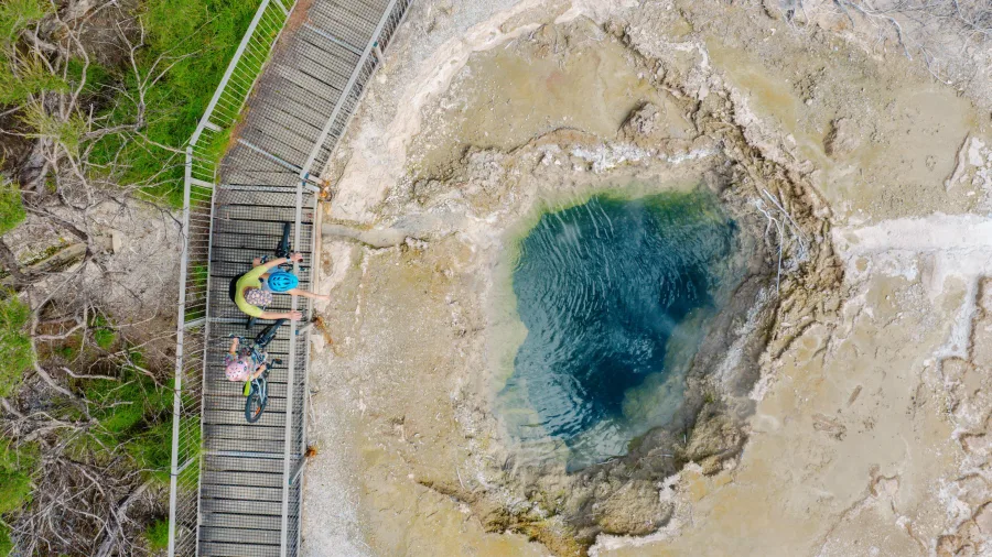 Overhead view of cyclists on a boardwalk looking down at a steaming geothermal pool along the Supervolcanic Cycle Trail in Turangi, New Zealand.