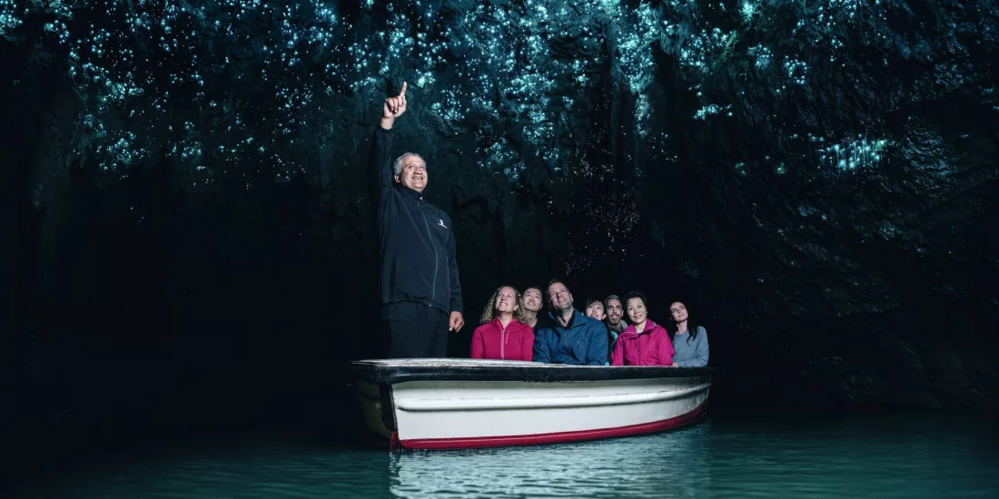 Visitors on a silent boat ride through the Waitomo Glowworm Caves with a guide pointing to the glowing cave ceiling