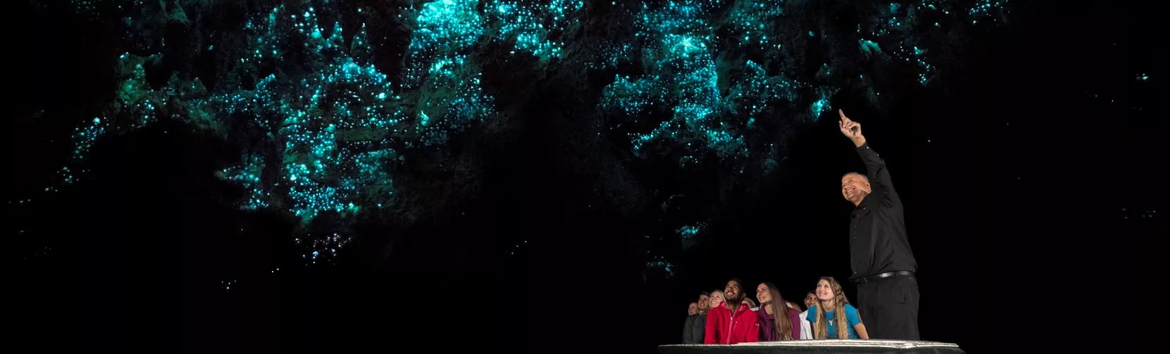 A small tour group in a boat gazing up at glowworms in the Waitomo Caves with a guide explaining the natural phenomenon