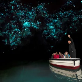 A small tour group in a boat gazing up at glowworms in the Waitomo Caves with a guide explaining the natural phenomenon