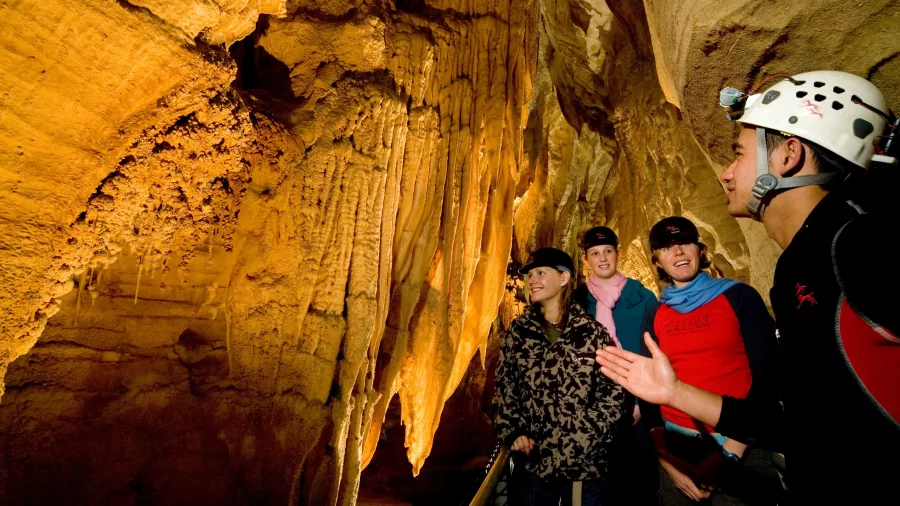 Waitomo guide explaining cave formations to a group of visitors wearing helmets inside an illuminated limestone cave