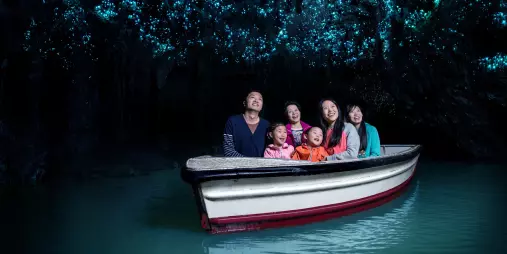 Family on a boat gliding through the Waitomo Glowworm Caves, lit by thousands of tiny blue glowworms overhead