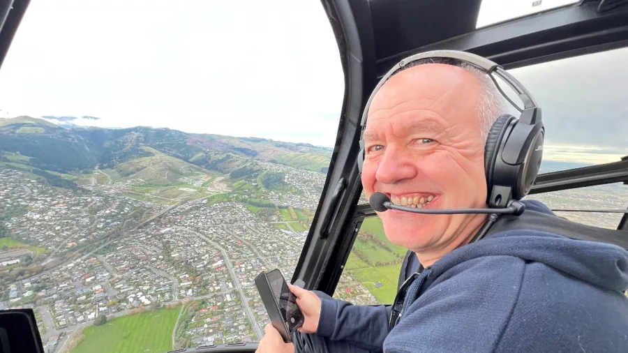 Man smiling inside a helicopter with aerial view of Wellington hills and suburbs in the background