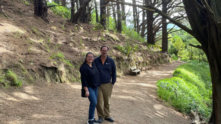 Two visitors standing at the filming location of the Nazgûl escape scene in Lord of the Rings, surrounded by pine trees