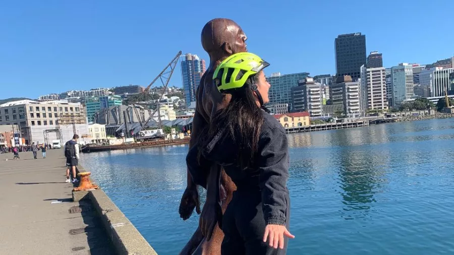 Woman in helmet posing playfully with the Leaning Man sculpture on the Wellington waterfront