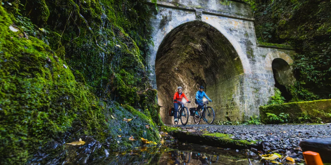 Two cyclists ride past a mossy rail tunnel entrance on the Remutaka Cycle Trail