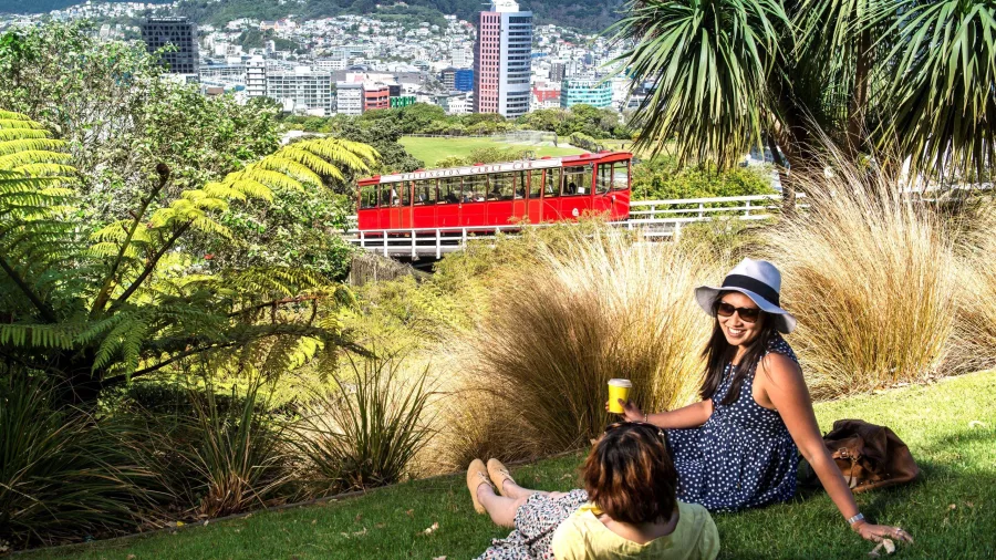 Two women relaxing on the grass with a view of the Wellington Cable Car passing by the Botanic Garden