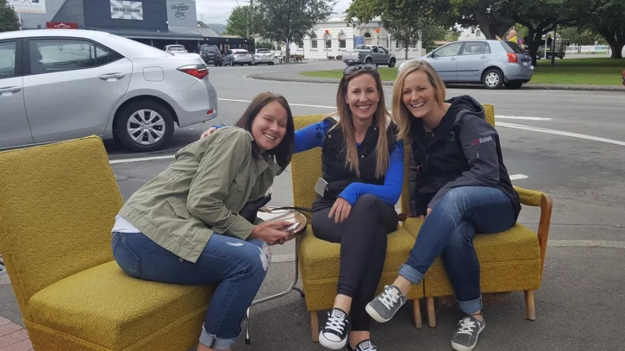Three women sitting on mustard yellow chairs on a street corner in Martinborough