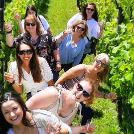Group of women smiling with wine glasses between vineyard rows in Martinborough