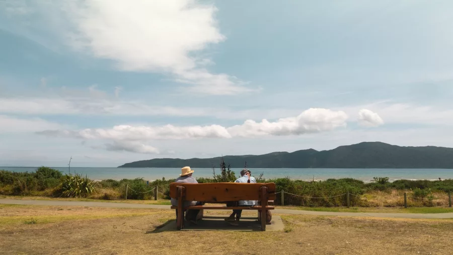 Two people sitting on a bench facing Kapiti Island from Paraparaumu Beach on a sunny day