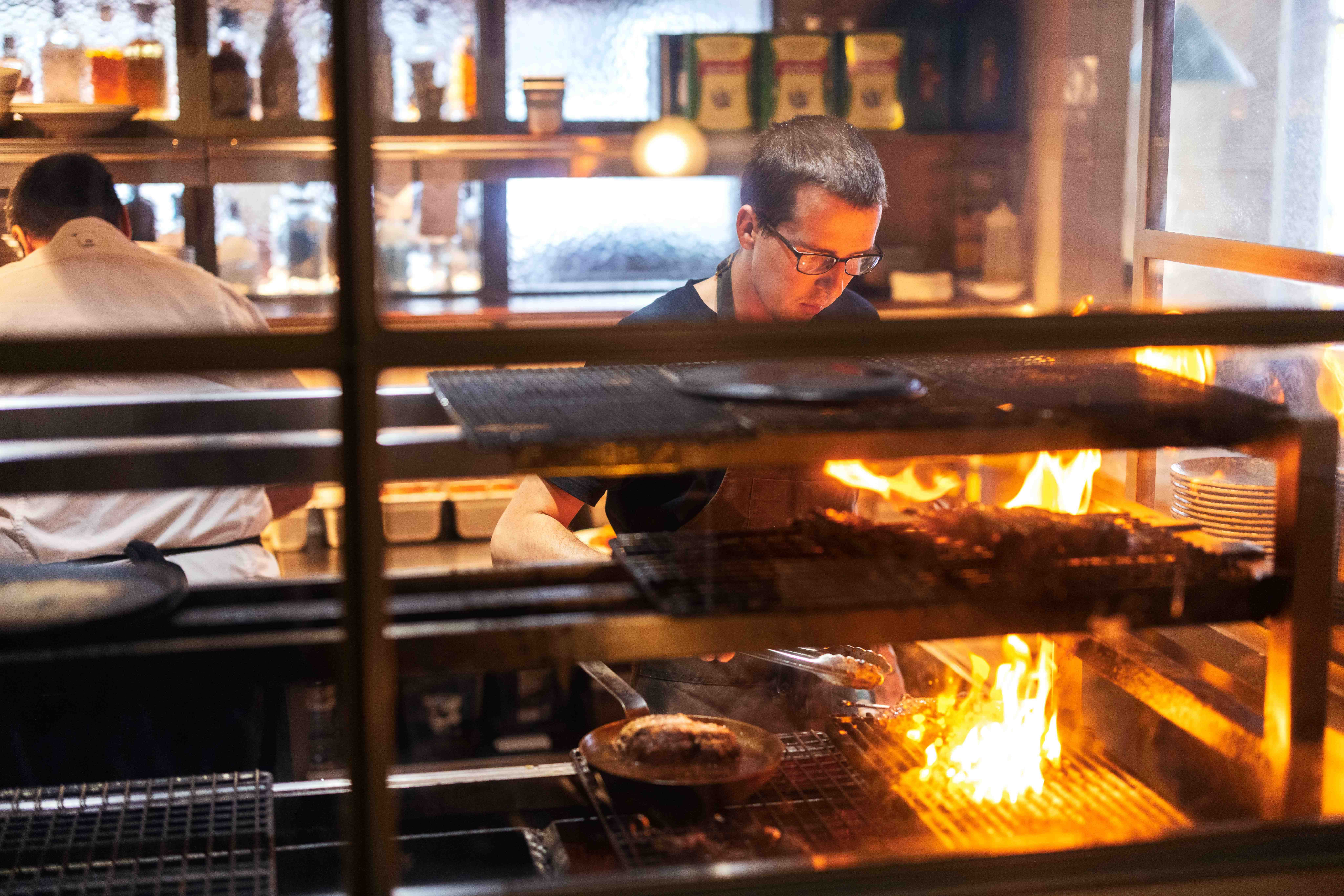 A chef cooking over an open flame in a restaurant kitchen in Te Aro, Wellington, New Zealand, seen through a window with warm firelight, shelves, plates, and kitchen equipment in the background. Photo by Miles Holden.