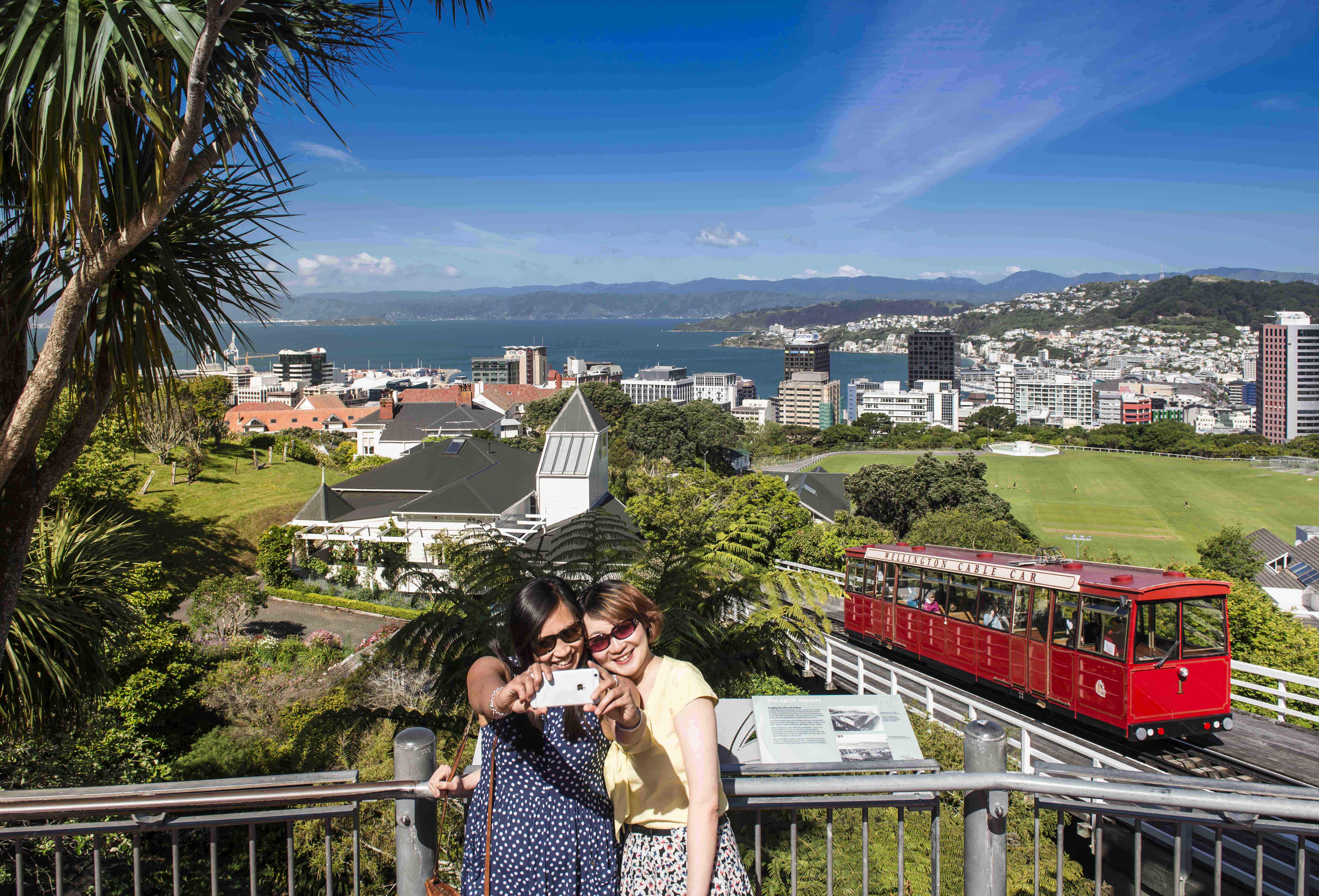 People are taking a selfie from the lookout at the top of the Wellington Cable Car with a city and harbour view on a summers day.