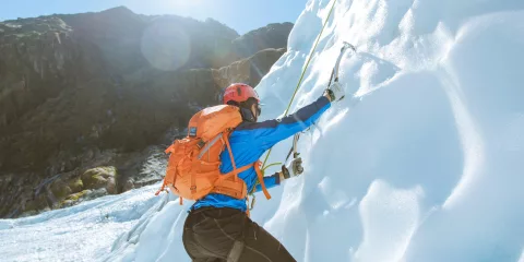 A climber uses ice axes to scale a frozen wall on Fox Glacier in bright alpine light.