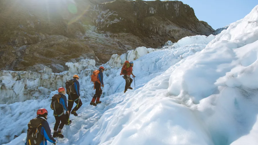 A line of hikers climbs the glacier with sunlight beaming over surrounding peaks.