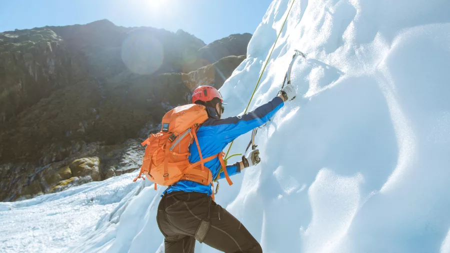 A climber uses ice axes to scale a frozen wall on Fox Glacier in bright alpine light.