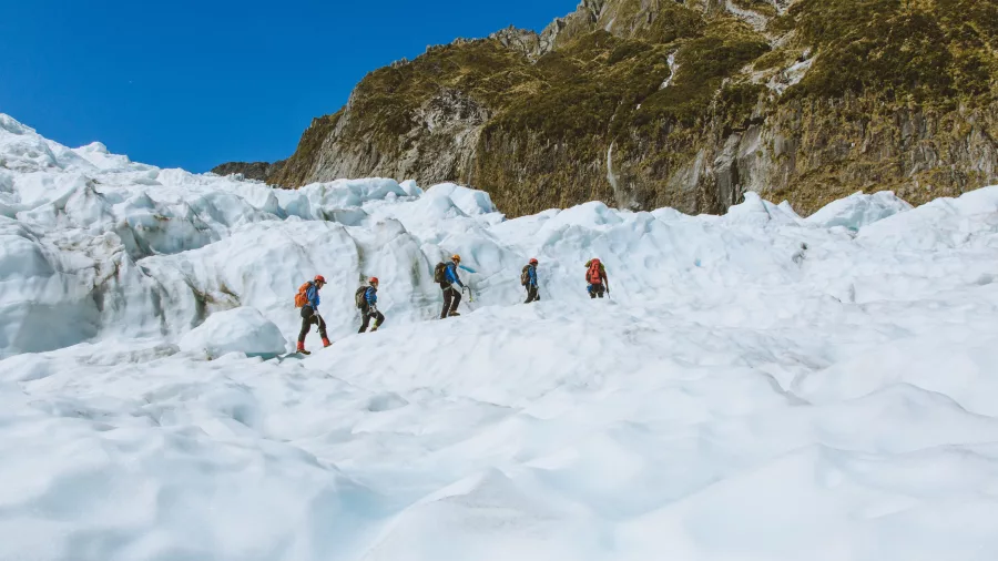 A team of hikers ascends a wide icy slope on Fox Glacier surrounded by rugged peaks.