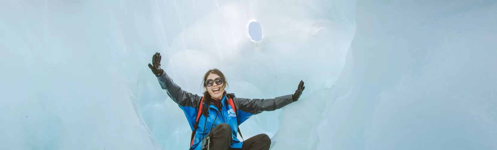 A smiling hiker sits inside a natural ice chamber on Fox Glacier.