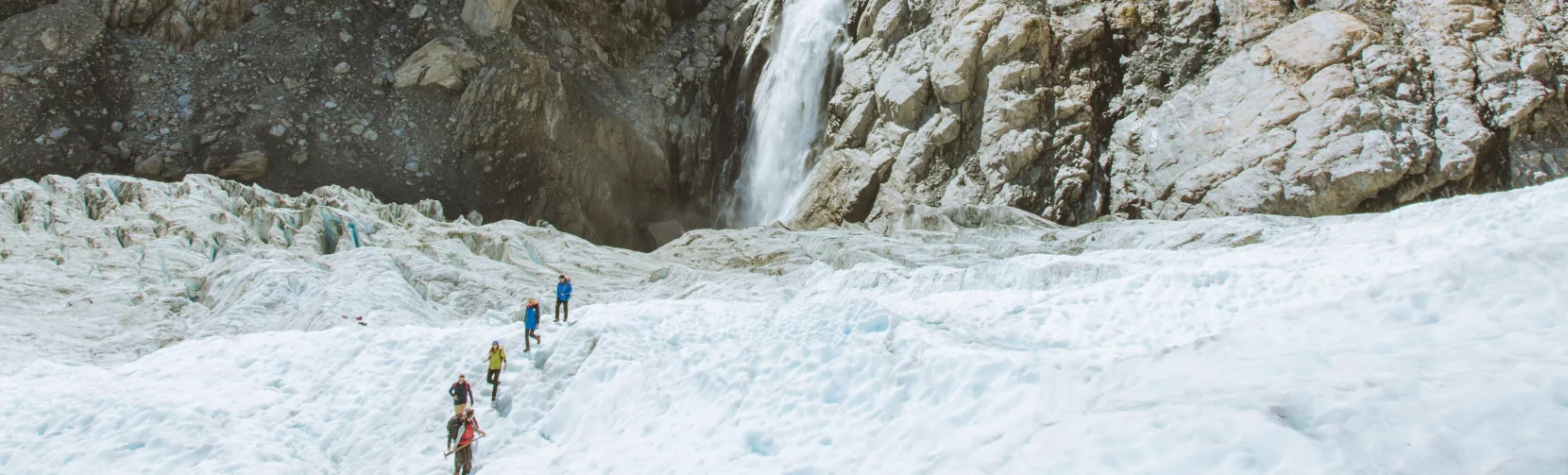 Hikers walk across the glacier with a towering mountain waterfall in view.