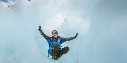 A smiling hiker sits inside a natural ice chamber on Fox Glacier.
