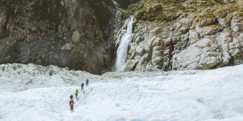 Hikers walk across the glacier with a towering mountain waterfall in view.